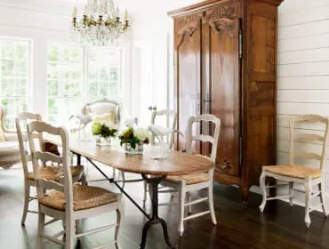 Dining room with a wooden table, upholstered chairs, a large armoire, and a crystal chandelier. White shiplap walls and large windows create a bright atmosphere.