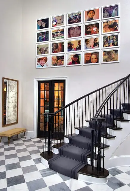 Entryway with a checkered marble floor, a dark wood staircase with a gray runner and black spindles, a mirrored wall hanging, and a grid of framed family photos on the wall.