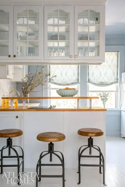 White kitchen island with wood countertop, three industrial-style stools, and glass-front cabinets displaying dishes and glassware.
