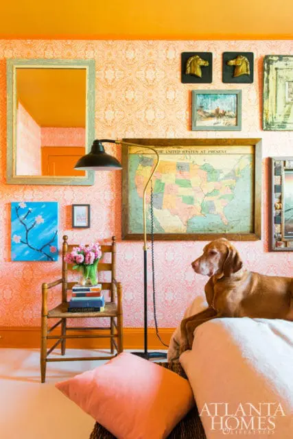 Orange patterned wallpaper adorns the walls of a room with a vintage wooden chair, a map of the US, and a dog resting on a bed.