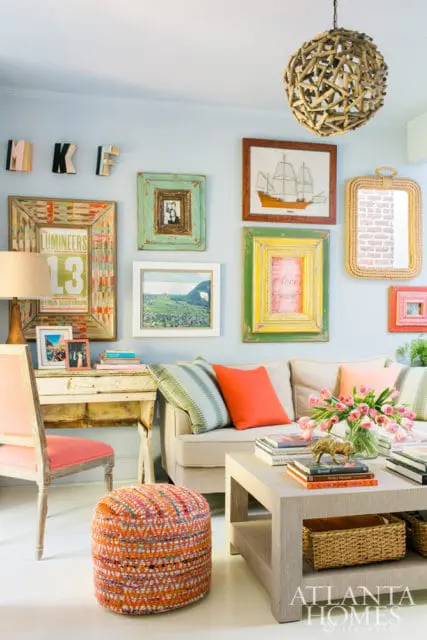 Light blue living room with a beige sofa, colorful gallery wall featuring framed art and photos, a distressed desk with a lamp, and an orange woven ottoman.