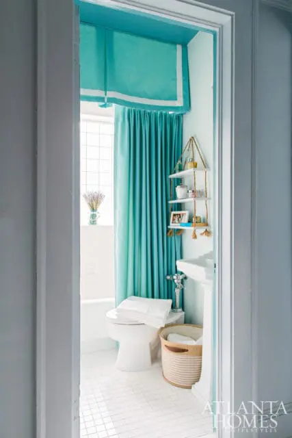 Small bathroom with white tile floor, white toilet with stacked towels, and teal curtains. A pedestal sink and hanging shelves with toiletries are visible.