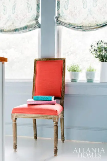 French country chair with coral upholstered seat and backrest, topped with two books on a light blue wall with patterned Roman shades.