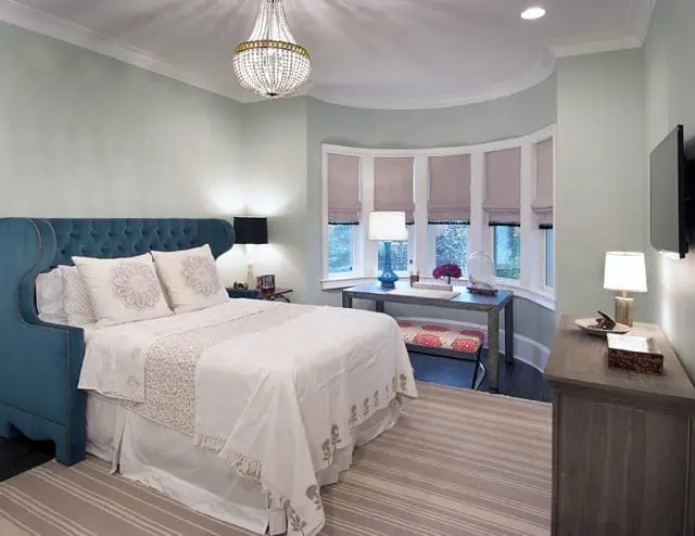 Bedroom with a teal tufted headboard, white bedding, a desk in a bay window, and a dresser with a television mounted above it.