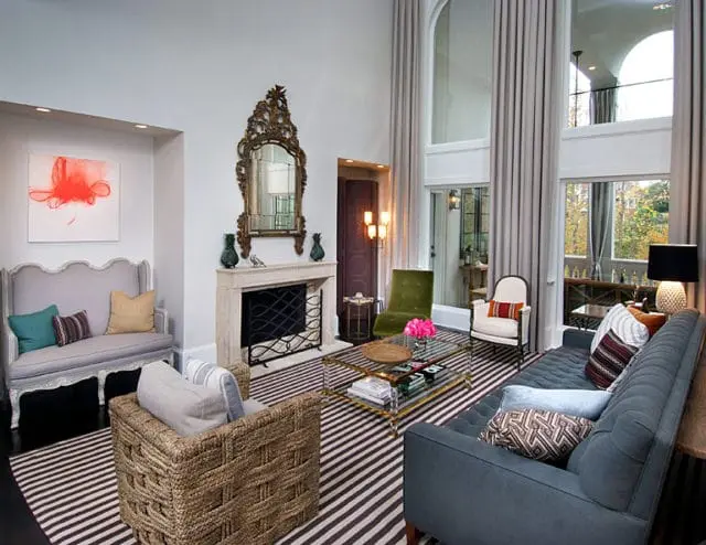 Living room with high ceilings, large windows draped in grey curtains, a marble fireplace with an ornate mirror, and a striped rug. Furniture includes a grey tufted sofa, a woven armchair, and a glass-top coffee table.