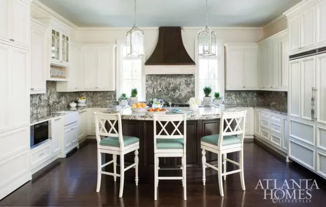 White kitchen with dark wood island, marble backsplash, and three white bar stools with teal seats.