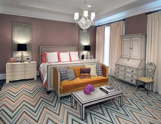 Master bedroom with a king-size bed, orange velvet sofa, chevron rug, and antique secretary desk. Walls are a muted lilac with distressed mirror panels.
