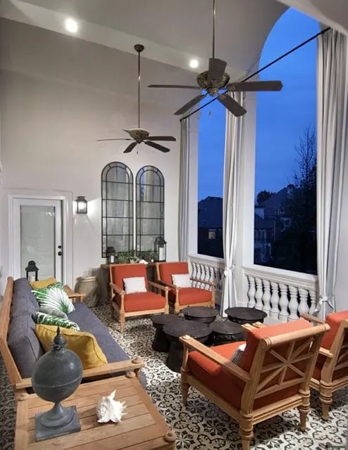 Covered patio with patterned tile floor, wooden furniture including a sofa and chairs with orange cushions, dark round tables, and ceiling fans.