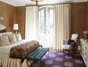 Bedroom with brown grasscloth walls, a cream upholstered headboard, and a colorful patterned bed runner. A tufted teal bench sits on a purple rug with white floral motifs.