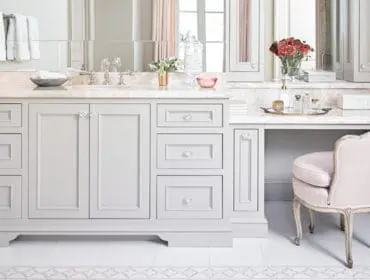 Light gray bathroom vanity with crystal knobs and a pink upholstered vanity chair. The vanity has a marble countertop and a large mirror above.