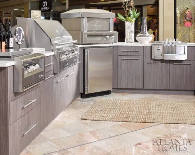 Outdoor kitchen with stainless steel grill and pizza oven, gray wood-grain cabinetry, and marble countertops.