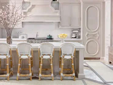 Cream kitchen with white rattan bar stools, marble countertops, stainless steel range, and decorative cabinetry.