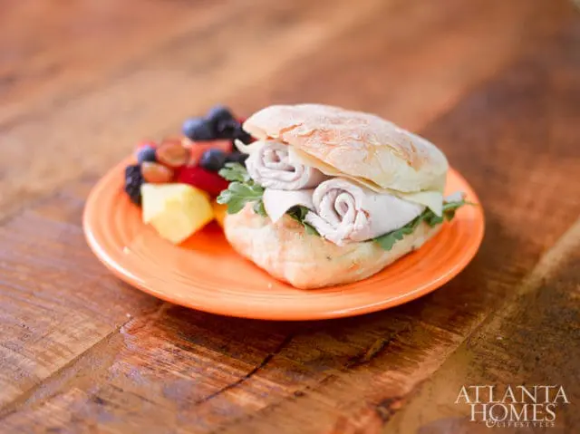 Close-up of a turkey sandwich on a ciabatta roll with cheese and arugula, served with a side of mixed fruit on an orange plate, resting on a wooden table.