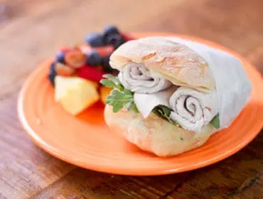 Turkey sandwich on a ciabatta roll with arugula, served with a side of mixed fruit on an orange plate