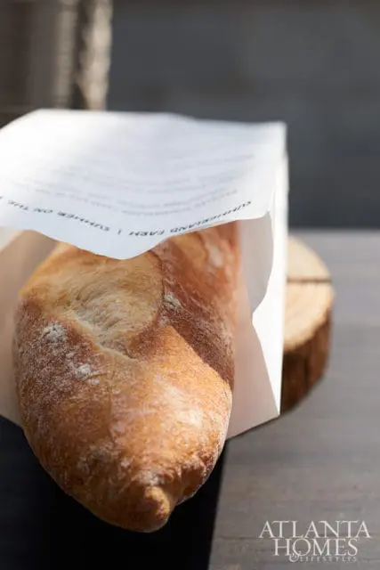 Close-up of a crusty baguette in a paper bag with text visible, next to a wooden slice.