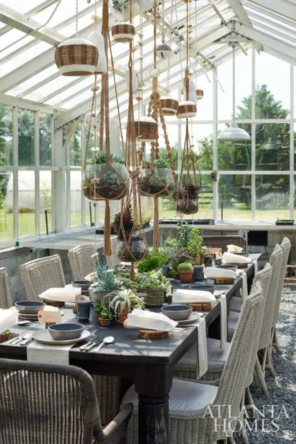 Greenhouse interior with a long dark wood dining table set for a meal, surrounded by wicker chairs. Macrame hangers with plants and baskets hang from the ceiling.