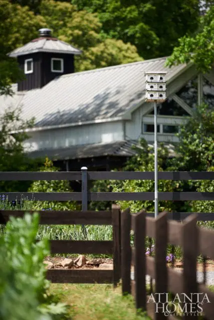 Modern barn with a metal roof and dark wood accents, featuring a multi-level birdhouse on a pole in front of a dark wooden fence.