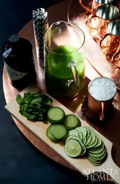 A bottle of Hendrick's Gin, a pitcher of green liquid, a glass of ice, a wooden cutting board with mint, cucumber slices, and lime slices, and copper mugs on a copper tray.