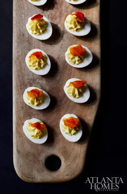 Deviled eggs with yellow filling and orange garnish on a wooden cutting board, with Atlanta Homes & Lifestyles logo in the corner
