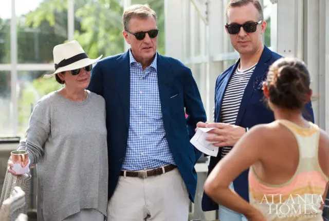 Three people in sunglasses stand together indoors. A woman in a grey sweater and white hat is on the left. A man in a blue gingham shirt and navy blazer is in the center. Another man in a striped shirt and navy blazer is on the right.