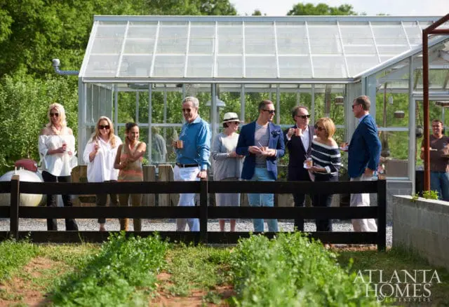 Group of people standing in front of a greenhouse, with a wooden fence in the foreground and lush greenery surrounding the area.