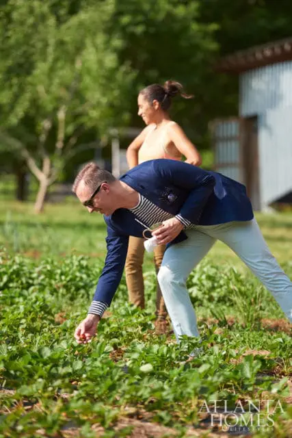 Man in navy blazer and light blue pants picking strawberries in a field, with a woman in a peach tank top standing behind him.