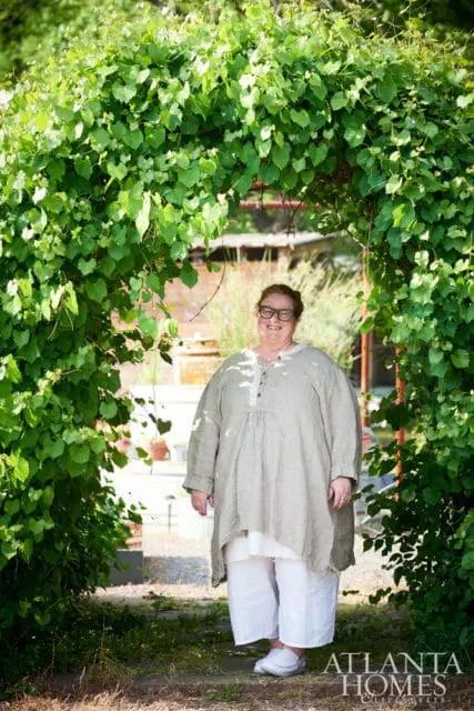 Woman in a linen tunic and white pants standing under a green vine-covered archway, with the Atlanta Homes & Lifestyles logo in the lower right corner.