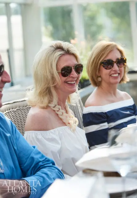 Two women wearing sunglasses and off-the-shoulder tops smile while seated at a table. A man in a blue shirt is visible on the left. The Atlanta Homes & Lifestyles logo is in the lower left corner.