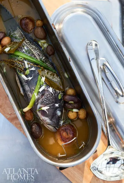 Whole fish cooked in broth with potatoes, mushrooms, herbs, and edible flowers, served in a metal pan with a ladle.