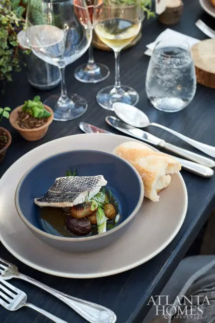 Close-up of a fish dish with potatoes and greens in a blue bowl, served with bread and wine glasses on a dark wooden table. Atlanta Homes & Lifestyles logo in the corner.