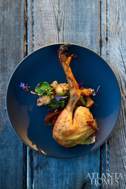 Roasted chicken leg and thigh served on a dark blue plate with a side of salad and edible flowers, set against a rustic wooden background.