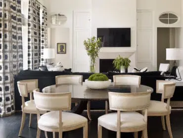 Dining area with dark wood table and upholstered chairs, adjacent to a living room with a black sofa, fireplace, and patterned curtains.