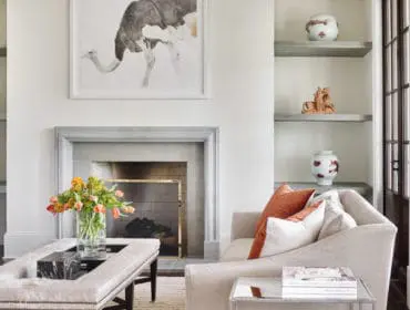 Living room with a fireplace, plush sofa with rust-colored pillows, and a tufted ottoman coffee table with a vase of flowers.