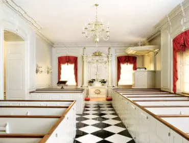 Church interior with white pews, red draped curtains, a black and white checkered floor, and a decorative altar with a cross.