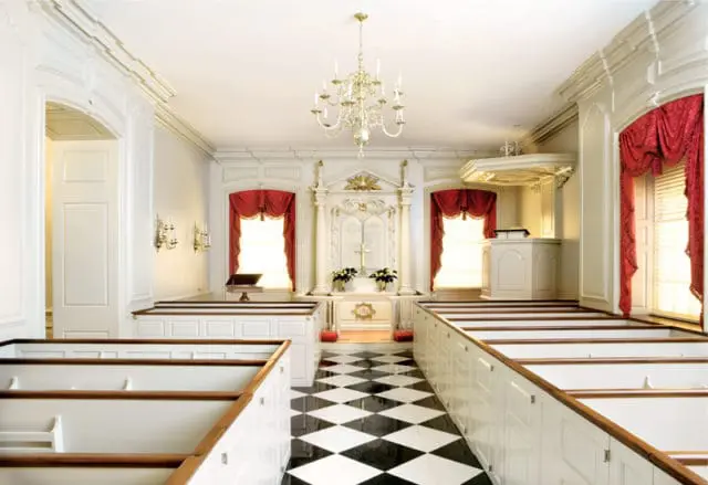Church interior with white pews, red draped curtains, a black and white checkered floor, and a decorative altar with a cross.