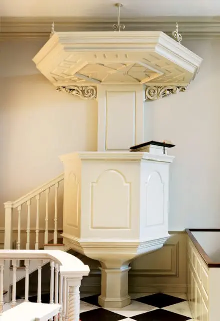 Cream-colored pulpit with ornate carved brackets and a coffered ceiling, set against a cream wall with a black and white checkered floor.