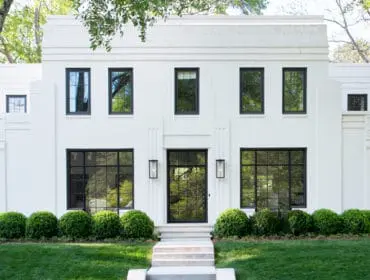 White Art Deco style house with black framed windows, a glass front door, and manicured boxwood bushes lining the front walkway.