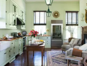 Light green kitchen with white farmhouse sink, wood countertops, stainless steel refrigerator, and a fireplace with two armchairs.