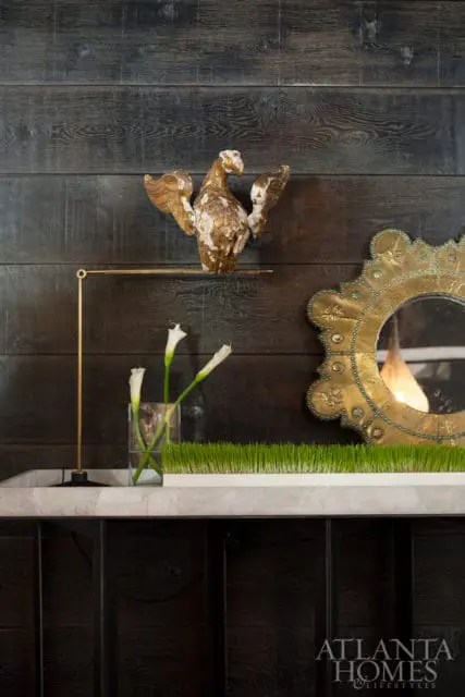 Dark wood paneled wall with a gold eagle sculpture, a brass lamp, white calla lilies in a glass vase, and a decorative gold mirror.