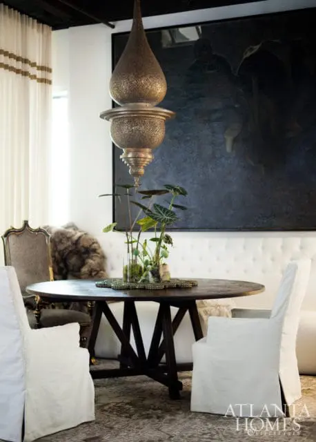 Dining room with dark wood round table, white slipcovered chairs, ornate chandelier, and large dark abstract painting on tufted white banquette.