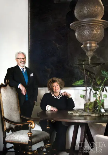 Man in a suit and woman in a black dress smiling at a table in a room with a large dark painting and ornate lamp.