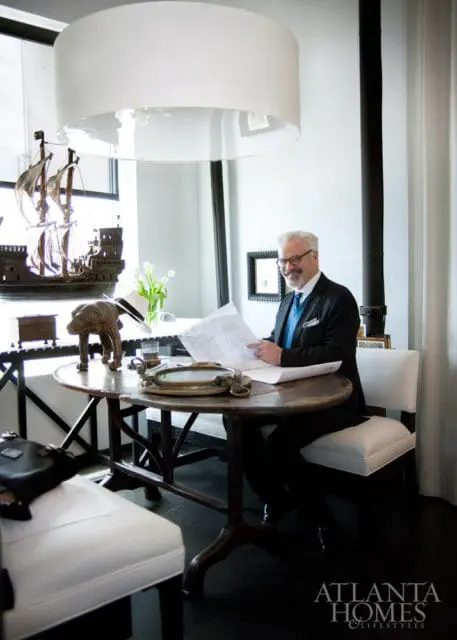 Man in suit reading blueprints at a round wooden table with a ship model and white tulips on display.