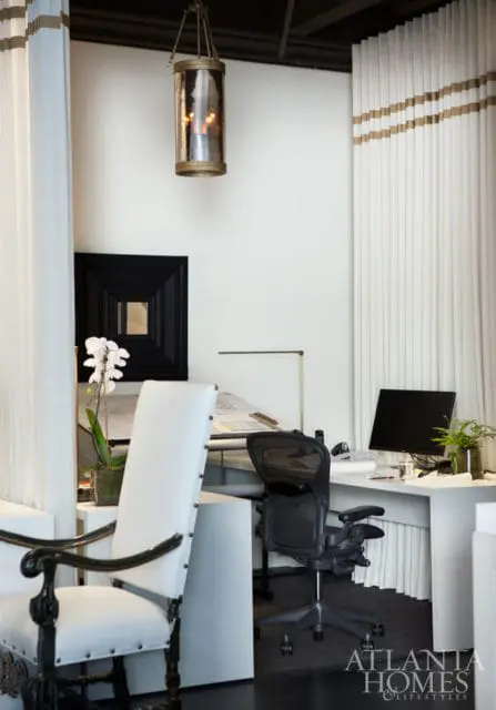 Modern office space with white walls, featuring a white drafting table with blueprints, a black ergonomic chair, and a computer monitor on a white desk. A decorative lantern hangs from the ceiling.