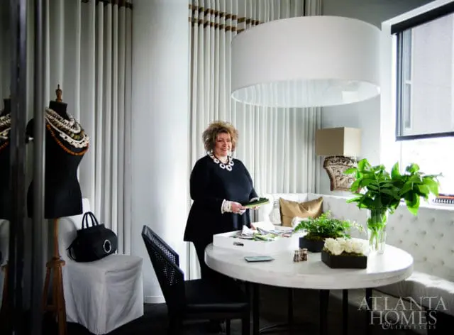 Woman in black dress standing by a round table in a room with white tufted banquette seating, floor-to-ceiling curtains, and a large white drum pendant light.