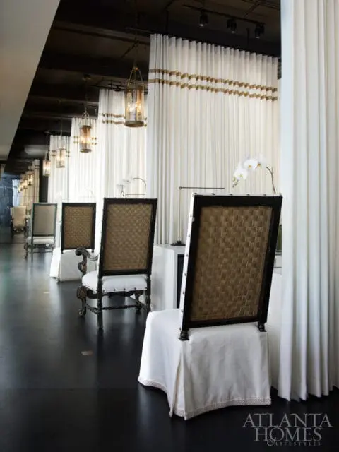 Elegant dining room with dark wood chairs featuring woven backs and white slipcovers, white drapes with gold trim, and hanging glass pendant lights.