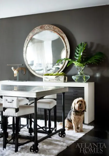 Dark gray walls with a large round distressed mirror above a white console table. A dog sits on a white rug next to a metal-framed table with white upholstered stools.