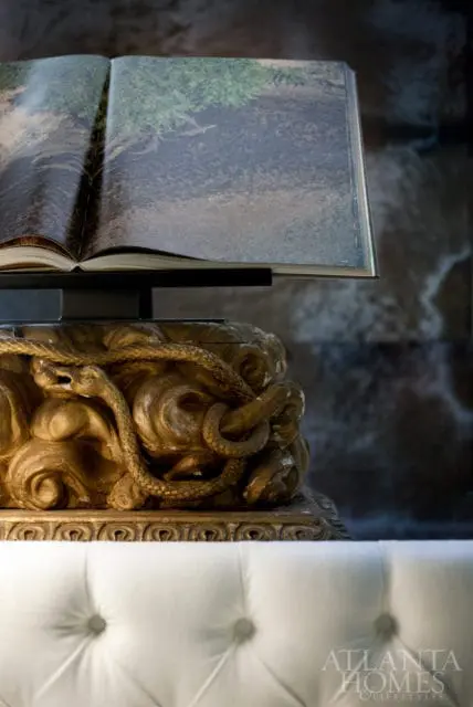 Open book displaying a landscape on a stand above a gold sculpture of a snake coiled around a face, with a tufted white sofa in the foreground.