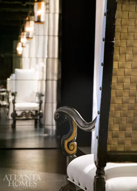Close-up of an ornate chair armrest with gold scroll detail, next to a white upholstered seat. In the background, a row of chairs and lanterns are reflected in a mirrored wall.
