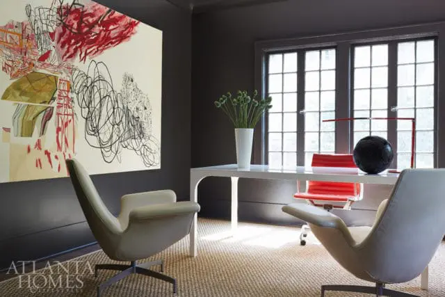 Modern office with dark gray walls, a white desk, two light gray chairs, and two red chairs. A large abstract painting hangs on the wall.
