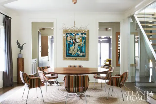 Dining room with a round wood table, striped upholstered chairs, abstract art, and a modern chandelier. A glass-paneled staircase is visible to the right.
